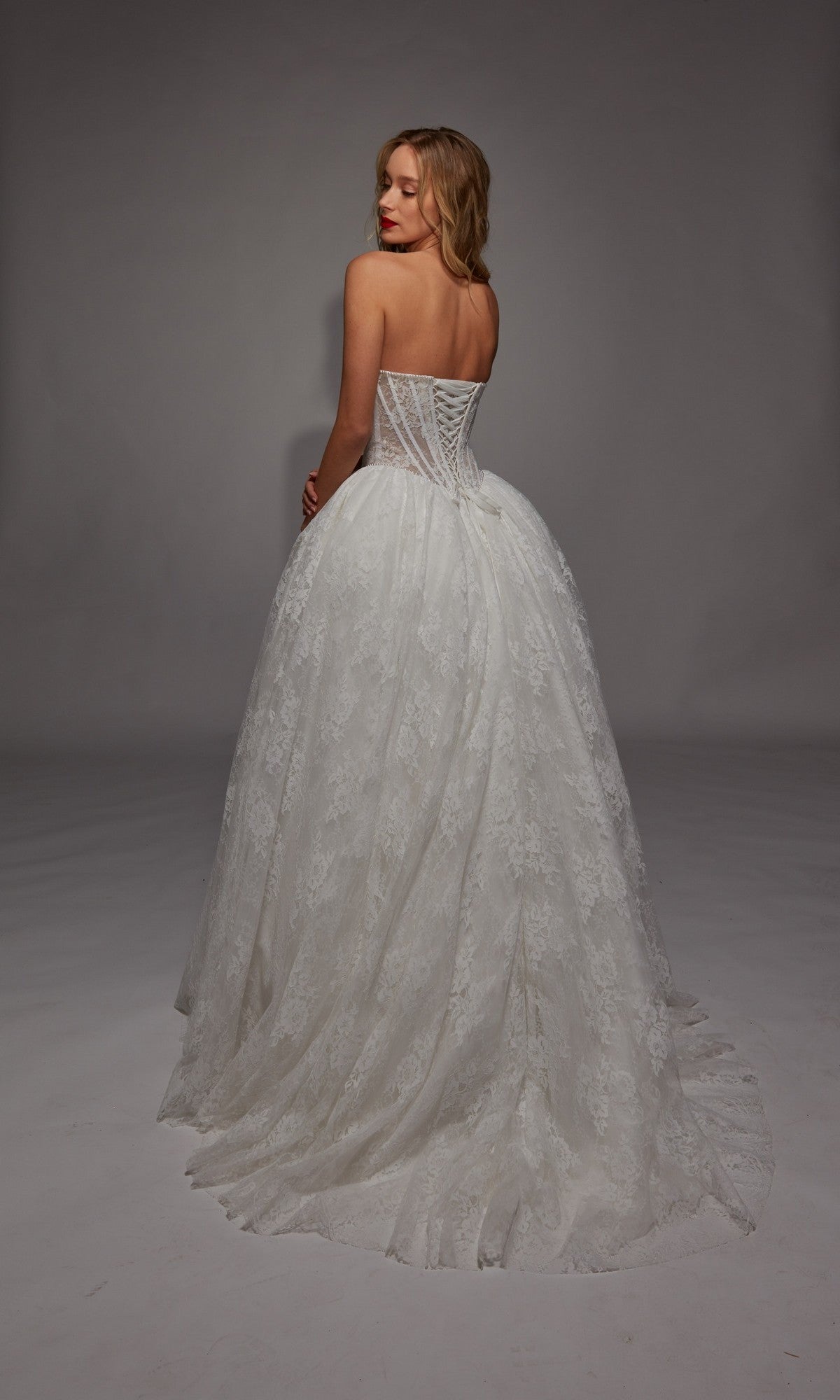A woman stands in a studio wearing the Alyce 7149, a long white strapless lace ball gown with a fitted corset back and flowing train—an elegant choice for a wedding dress.